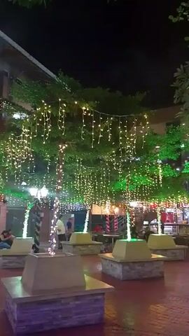 The video depicts an outdoor courtyard at night illuminated by numerous strings of light hanging from trees and lining the upper facade of a building. Several white square bench-like structures with stone bases are scattered around the brick-paved area, some with people sitting on them. A person wearing a white shirt walks past a store labeled "Auroda" in orange letters. The tree trunks are wrapped with white lights.