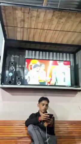 A man is sitting on a bench, looking at his phone. Above him, a television is mounted on the wall, displaying a soccer game. The TV shows players' legs and feet in soccer shoes. The man sneezes. There are speakers to the left of the television. The area appears to be a semi-outdoor seating space with a roof.