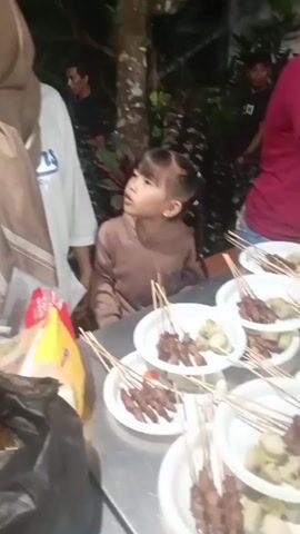 The video shows a group of people, including women and children, gathered around a table laden with plates of food. The food appears to be satay, with skewers of meat arranged on white paper plates alongside what look like rice cakes. In the foreground, a woman wearing a hijab and a white t-shirt stands to the left, while a young child in a brown outfit looks towards the table. To the right, an older woman in a red shirt and other individuals are visible, with the older woman appearing to serve or arrange the food. The setting appears to be outdoors, possibly a garden or backyard, with trees and foliage visible in the background, and the lighting suggests it is evening or nighttime. There is also a black plastic bag and some yellow packaging on the table.