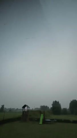 The video shows a cloudy sky over a backyard. In the foreground, there is a green lawn. In the middle ground, there is a wooden play structure with a slide and a trampoline. In the background, there are trees and a fence. The sky is overcast and appears to be raining lightly. The overall mood is gloomy.