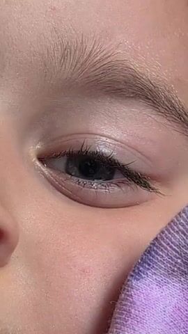 The video is a close-up of a person's face. It begins with a shot of the left eye and eyebrow, then pans down to show the lips and chin, and finally shows the eye, nose, and lips from a slightly different angle. The person has blonde hair and light-colored eyes. The background is blurred and a purple and white patterned fabric is visible on the right side of the frame.