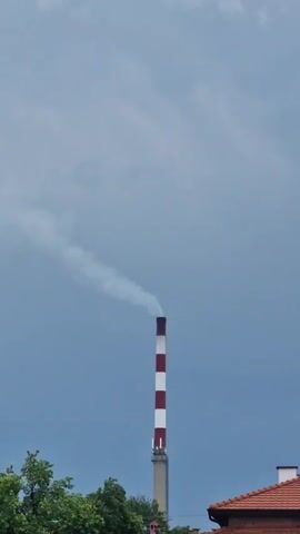 The video features a tall, red and white striped industrial chimney emitting a plume of white smoke or steam into an overcast sky. The smoke drifts horizontally to the left from the chimney's top. In the foreground, green treetops are visible at the bottom left. On the right, the orange-red tiled roof of a building can be seen. The overall scene suggests an industrial area under cloudy, daytime conditions. The location is Katunitsa, Bulgaria. No people or visible text are present.