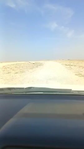 The video shows a first-person view from inside a car, looking out through the windshield. The car is driving on a dirt road in a desert landscape. The road is light-colored and appears to be made of sand and gravel. On either side of the road, the ground is covered in sand and sparse vegetation. The sky is blue with a few wispy clouds. The dashboard of the car is visible in the foreground, and the windshield wipers are also visible. The video appears to have been filmed during the day.