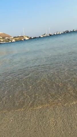 This is a short video clip of a beach scene. The foreground shows the sandy shore and the clear, shallow water of the sea. Gentle waves are breaking on the beach. In the background, there is a body of water with several boats and yachts anchored. Beyond the water, there is a coastline with hills and buildings, suggesting a populated area. The sky is clear and blue, indicating a sunny day. The overall atmosphere is peaceful and serene. The time of day appears to be daytime, possibly late afternoon, given the angle of the sunlight creating patterns on the water. There is no visible text in the video.