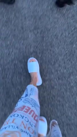 A first-person perspective video shows a person walking on a paved surface. The person is wearing white slippers and pants with an orange and white pattern. In their hands, they are holding two white objects with orange tips. Another person is partially visible in the upper left corner of the frame, wearing dark footwear. The environment appears to be outdoors, with the ground being asphalt or a similar material.