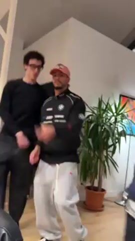 Inside a bright, modern living space, two men are dancing energetically to music. The first man, wearing a black long-sleeved shirt, glasses, and black pants, dances with fluid, arm-centric motions. The second man, in a black BMW-branded sweatshirt, light grey sweatpants, and a pink baseball cap, initially faces the camera, gesturing and moving before turning to dance with the first man. A large potted plant sits near the second man, and a painting hangs on the wall. The room features a high ceiling with exposed beams and track lighting.