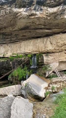 A video shows a small waterfall cascading over mossy rocks at the base of a large rock overhang. Lush green vegetation grows around the rocks and on the overhang. In the background, a steep, rocky hillside covered in greenery is visible. Several large boulders are scattered in the foreground. A ladder leans against the rock face to the right of the waterfall. The overall scene depicts a natural, somewhat rugged landscape.