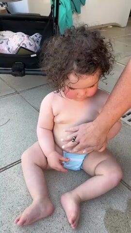 A baby wearing a blue and white striped diaper is sitting on a tiled patio. The baby has curly brown hair and rosy cheeks. A person's hands are visible near the baby's diaper and leg. In the background, a stroller with a pink and white patterned item is visible. The baby looks towards the right of the frame and then turns its head to look up and to the left.