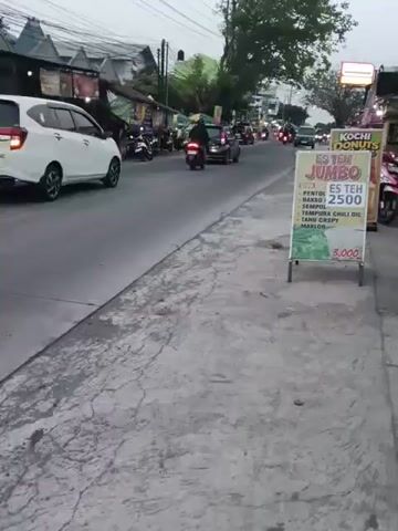 This video captures a street scene, likely in Southeast Asia, given the writing on the signs. The time of day appears to be late afternoon or early evening, with overcast skies. The camera is positioned on a sidewalk, looking towards a busy road.

On the road, vehicles, primarily motorcycles and cars, are moving in both directions. Several motorcycles are visible, with riders wearing helmets. A white MPV is in the left lane, and other cars are further down the road.

Along the right side of the road, there are several storefronts and food stalls. One prominent sign displays an advertisement for "ES TEH JUMBO" (Jumbo Iced Tea) with a price of 2500. Below this, it lists ingredients or accompaniments like "PENTOL," "BAKSO," "TEMPURA CHILI OIL," and "MAKLON." Another sign is for "KOCHI DONUTS." A pink scooter is parked near the donuts stall.

The sidewalk where the camera is located is cracked and worn. There are some potted plants and a stand holding the advertisement for iced tea. The overall environment suggests a commercial or residential area with moderate foot traffic and vehicular activity.