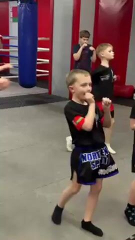 In a gym, a young, fair-skinned boy in black athletic shorts that say “Northern Spirit” and a black t-shirt is practicing martial arts moves with a group of other children. Behind him, a boxing ring with red ropes and a blue punching bag are visible. The floor is grey with black lines. The boy practices kicks and hand movements, touching his finger to his face several times. Other children in similar clothing move behind him, mirroring the motions.