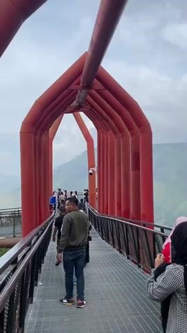 The video opens with a medium shot of a modern red arched walkway against a cloudy sky. The walkway, made of metal, has a patterned railing on either side and a wide, flat surface. Several people are walking along the walkway. In the foreground, a man in a dark jacket and jeans walks away from the camera. A woman wearing a pink hijab and red jacket appears in the lower right corner and gestures towards the view. In the background, rolling green hills are visible beyond the walkway's edge. The camera pans slightly to the left, revealing more of the walkway and the people on it, as well as a security camera mounted on one of the red arch supports. The overall scene appears to be an outdoor tourist attraction with scenic views.