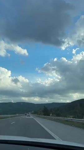 The video shows a view from the inside of a car as it travels on a highway. The car's dashboard is visible in the foreground, and the road stretches out ahead, lined with guardrails and a grassy shoulder. Several other cars are also on the highway, moving in the same direction. The landscape on either side of the road consists of rolling hills covered in trees. The sky above is a mix of blue sky and dramatic clouds, some of which are dark and stormy, while others are bright and puffy. A contrail from an airplane is visible in the distance. The lighting suggests it is daytime, possibly late afternoon or early evening.