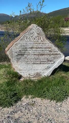 The video shows a landscape with a river and a stone monument. The monument, labeled "Колвица" (Kolvitsa), is inscribed with text in Russian and sits amidst green grass and gravel. Behind the monument, a river flows through the landscape, with a sandy bank visible on the opposite side. The sky is a clear blue, and distant mountains are visible. The camera pans from the monument to the river and the landscape.