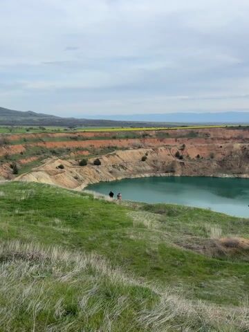 The video presents a scenic view of an open-pit mine filled with striking turquoise water in Tsar Asen, Bulgaria. The camera initially captures a wide shot of the landscape, showing two individuals standing on the grassy edge of the pit, looking down at the water. The pit's walls are terraced, revealing layers of reddish-brown and tan rock and soil. A small, dark green tree stands near the water's edge within the pit.

Beyond the mine, the landscape extends into rolling hills with patches of green and some fields showing bright yellow blooms, possibly rapeseed, in the distance. Further back, mountains are visible under an overcast sky. As the video progresses, the camera slowly zooms in, offering a closer look at the vibrant turquoise water and the rugged texture of the mine walls. The foreground is dominated by green, slightly windswept grass. The overall atmosphere is calm and observational, focusing on the natural beauty and human alteration of the landscape.