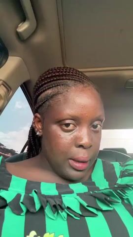 A woman with dark skin and braided hair sits in the driver's seat of a car. She is wearing a green and black striped shirt with ruffles. Her expression is serious. The car has a sunroof and leather seats. The sky is visible through the window.