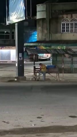 A person wearing a yellow shirt and blue pants sits at a wooden stall by the side of a street at night. The stall has a metal roof and appears to be a food vendor. The person is hunched over, possibly looking at a phone or preparing food. The street is paved and appears mostly empty. In the background, there are buildings, some with lights on, and a large billboard. The overall atmosphere is dimly lit and quiet.