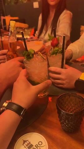 The video shows a group of four people toasting drinks together at a wooden table in what appears to be a bar or restaurant. Four hands holding glasses filled with ice and garnished with fruit and mint are prominently visible in the foreground. Black straws are inserted in the drinks. One person is wearing a watch with a black leather strap, another is wearing a watch with a yellow band. There is a dark, patterned glass next to the drinks. Behind the hands, the torsos and faces of three people are partially visible. One is a woman with long dark hair wearing a white top, taking a picture with her phone. A bottle of beer with a lime wedge is also on the table. The table is decorated with red and orange accents, and there is a coaster with an image of a pagoda on it. The background includes blurred foliage. The lighting is dim and warm, suggesting an intimate and relaxed atmosphere.