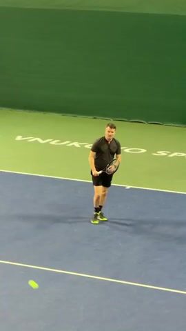 A male tennis player is featured on an indoor tennis court. He wears a dark grey t-shirt, black shorts, and black and lime green athletic shoes. The court is blue with white lines, and there's a green area beyond the baseline with partially visible white text that appears to read "NUKOVO SPORTS".

The video shows the player in two sequences. Initially, he hits a forehand stroke from the baseline, displaying a full swing and follow-through. He then walks towards the net area, bending down to pick up a tennis ball. In the second sequence, he stands near the net, bouncing the tennis ball with his hand while holding his racket, appearing to prepare for a serve or practice. The lighting is bright and consistent, typical of an indoor sports facility.