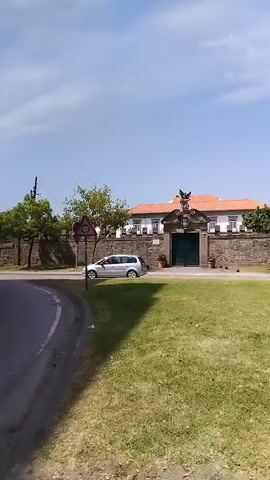 The video pans across a street view with a building in the background. The building has an orange tiled roof and a stone wall with a large ornate gate. There is a large green lawn in front of the building and trees on either side. A white van is parked on the side of the road. Traffic signs are visible on the side of the road. The sky is blue with some clouds. The camera is moving to the right as the video progresses.