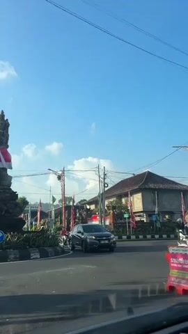 The video begins facing a stone monument adorned with red and white fabric, situated in the center of a traffic circle. A blue directional arrow sign is visible to the left of the monument. The background shows a clear blue sky with scattered clouds, telephone wires, trees, and buildings. A black car is positioned to the right of the monument, partially obscured. As the video progresses, the camera pans slightly to the right. A person on a scooter, wearing a brown jacket and helmet, is seen riding away from the camera towards the left side of the frame. Further down the road, more people on scooters are visible, along with flags and signs on the sides of the street, suggesting a populated area, possibly a town or village. The road appears to be paved, and there are trees and houses lining it. The overall environment suggests a sunny day in an urban or suburban setting, likely in a tropical or subtropical region given the vegetation.