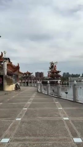 The video shows a walkway near Lotus Pond in Kaohsiung, Taiwan. The sky is overcast. The walkway is paved with gray tiles and bordered by a white railing. Statues of deities stand on pavilions in the water. Buildings are visible in the background. People are walking along the walkway. A bicycle is parked near a building at the edge of the frame.