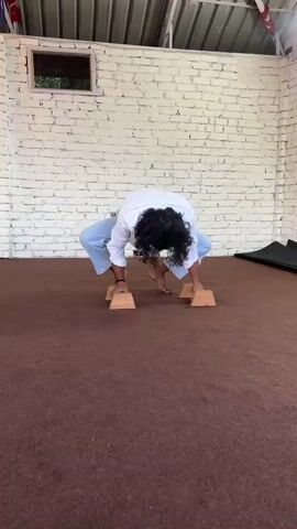 A person with long dark curly hair wearing a white long-sleeved shirt and light blue pants is performing a handstand exercise. They are balanced on two wooden blocks placed on a brown carpeted floor. The person's body is inverted, with their hands on the blocks and their legs spread wide in a straddle position. The background features a white brick wall and a corrugated metal ceiling.