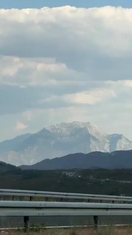 The video shows a scenic view of mountains and clouds from the perspective of a moving vehicle. A road guardrail is visible in the foreground. In the middle ground, there are rolling hills covered in green vegetation, and several wind turbines are scattered across the landscape. In the background, a large, rugged mountain range is visible, with its peaks appearing somewhat hazy due to the distance and atmospheric conditions. A black car drives by on the road in the foreground. The sky is partly cloudy with bright white cumulus clouds against a pale blue sky.