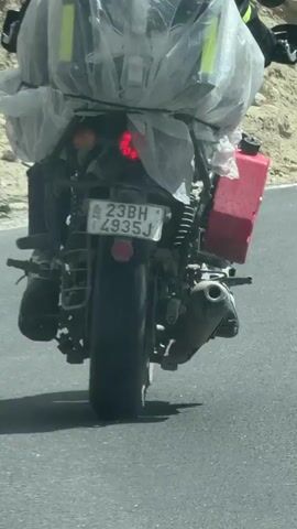 The video shows a person riding a motorcycle loaded with luggage on a paved road in a mountainous region. The camera perspective shifts from behind the motorcycle, showing the rider and their gear, to a view from inside a car following the motorcycle. The landscape is characterized by rugged, barren mountains and a clear sky with scattered clouds. The road is asphalt with white markings. Power lines are visible along the roadside, and there are some rocks and dirt on the slopes.