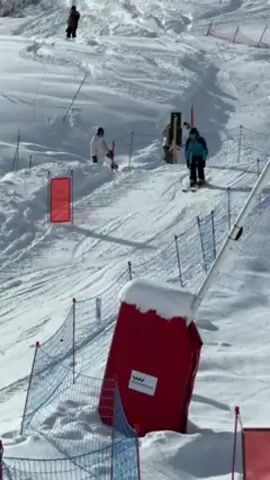 The video shows a person skiing on a snowy slope. The skier approaches a small jump, launches into the air, and performs a backflip, landing successfully. The scene takes place in a winter environment with snow-covered mountains and trees in the background. A building with the word "PLATTIER" written on it is visible nearby.