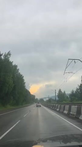 A dashcam view of a wet, two-lane road passes by trees and a railway line on a cloudy day. A white car is visible ahead, and power lines run along the right side of the frame.