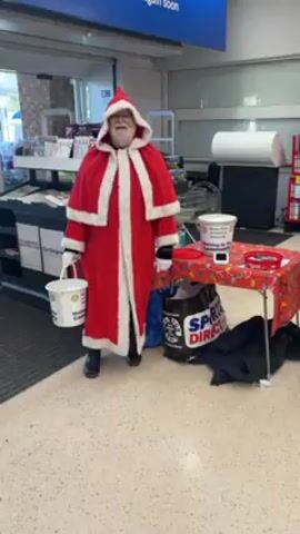 The video shows a person dressed as Santa Claus standing in front of a table with a collection bucket at what appears to be the entrance to a store. Another woman stands next to the table; she is wearing a vest with the logo of a charitable organization. A table sits between them, covered with a Christmas-themed cloth. There is a collection bucket on the table, as well as bowls with what look like candies. Bags are also visible under the table. Customers walk past them with their shopping carts.  The interior of the store is visible behind them.