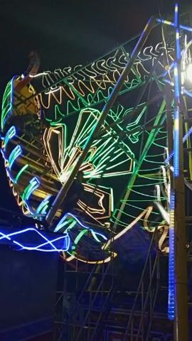 The video shows a rotating amusement park ride at night. The ride is decorated with neon lights in green, yellow, and blue. The camera is positioned close to the ride, and the perspective shifts slightly as it rotates. In the background, there are support structures for the ride and a dimly lit area with some people visible in silhouette. The music is loud, featuring a combination of brass instruments and percussion. The overall impression is of a brightly lit, festive attraction at a nighttime fair or amusement park.