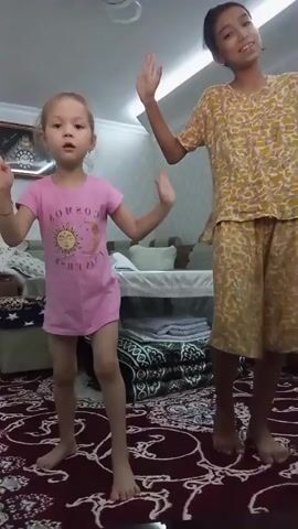 Two girls are dancing in a room. The girl on the left is younger, wearing a pink t-shirt with a sun graphic and text that reads "COSMO". She has blonde hair and is wearing no pants. The girl on the right is older, wearing a yellow patterned t-shirt and matching shorts. She has dark hair and is smiling. They are both barefoot. The room appears to be a living room, with a patterned rug on the floor and furniture in the background, including a couch and what appears to be a crib. There is also a light fixture on the ceiling. The overall mood seems happy and playful. The image quality suggests it was taken indoors during the daytime.