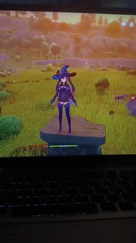 The video shows a laptop with a video game displayed on its screen. The game appears to be Genshin Impact, featuring a character named Mona standing on a stone platform in a grassy field. The character is wearing a blue and purple witch's outfit. The background depicts rolling hills and a sky with warm, sunset-like colors. The laptop's keyboard, with its illuminated keys, is visible in the foreground, out of focus. The overall scene is a snapshot of someone playing a video game.