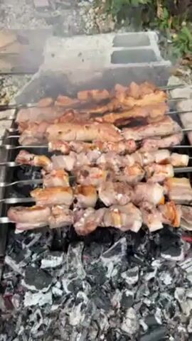 The video shows meat grilling over charcoal embers on metal skewers. Two types of meat are visible: skewers with chunks of meat and skewers with thin slices. Smoke rises from the coals and meat, and occasional flames appear between the coals. The grill is constructed with concrete blocks. The environment appears to be outdoors, with green plants visible at the edges of the frame. The video is taken from a slightly elevated angle.
