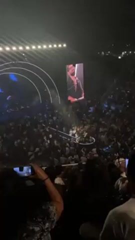 Here's a description of the video:
The video shows a large crowd of people in an arena setting, likely at a concert or event. A stage is visible in the center with a performer on it. A large screen above the stage displays a close-up of the performer, who appears to be singing or speaking into a microphone. Many audience members are holding up their phones, seemingly recording or taking pictures of the performance. Lighting and stage design elements are visible in the background. The video is taken from a high vantage point within the audience.