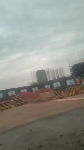 The video shows a view from a moving vehicle of a construction site on a cloudy day. There's a dark fence with white logos, piles of dirt, and yellow and black striped barriers. In the background, there's a structure covered with green netting and steel scaffolding. Power lines and poles are also visible. The road is gravelly and unpaved.