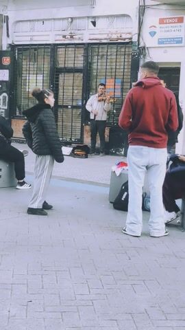 The video captures an outdoor street scene in Lanús Este, Argentina, during daytime hours. The footage is vertically oriented, giving the impression of the ground being a wall.

**Subjects:** Several individuals are present. A male street musician in a white shirt and dark pants is actively playing a violin. In the foreground, a person in a black puffer jacket and light grey sweatpants with white stripes stands observing the musician. Another person in a red hoodie and light-colored pants walks past, briefly stopping before continuing. A third person, wearing a blue puffer jacket and jeans, also walks across the frame. Other partial figures are visible in the background.

**Objects:** The musician uses a violin and bow. A black instrument case or bag is on the ground near him, and a black Puma brand bag is also visible nearby. A real estate "For Sale" sign is affixed to the building on the left.

**Scene/Setting:** The scene unfolds on a paved sidewalk, characterized by rectangular light-colored tiles. Buildings line one side of the street, featuring metal grates or a roll-down door behind the musician.

**Activity or event taking place:** A street musician is performing the violin, seemingly busking. Pedestrians are seen walking by, with some pausing briefly to listen or observe the performance.

**Notable details:** The lighting suggests an overcast but bright day. The overall mood appears casual, with people moving through their daily routines.

**Visible text:** The sign on the building clearly displays "VENDE" (For Sale), "MAURO MARVISI", contact numbers "11 5963-4745" and "4247-2529", and the real estate agency name "RE/MAX D+A HOUSE".