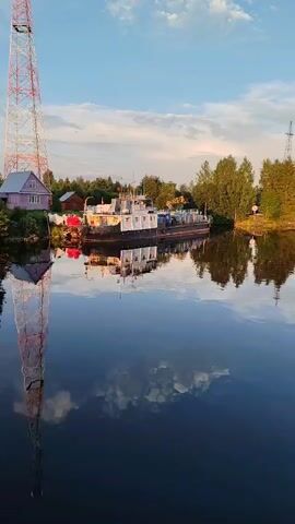 A vertical video shows a calm body of water reflecting the sky, clouds, trees, a red and white tower, a small house, and a docked boat. The boat appears to be a barge with a white superstructure and a dark hull. The tower is very tall and extends beyond the top of the frame. The scene is illuminated by what appears to be natural light, possibly from a sunrise or sunset, given the warm tones in the sky and the reflections. The overall atmosphere is peaceful and serene.