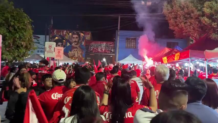 A large crowd of people wearing red shirts with the word "Corona" on the back are gathered in a street at night. There are tents set up on either side of the street, and a red flare is burning in the middle of the crowd, creating a large plume of white smoke. A mural is visible on a building behind the crowd. The crowd appears to be celebrating.