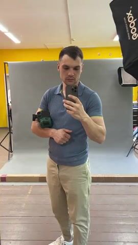 A man is taking a selfie in a mirror. He is wearing a blue collared shirt with yellow trim, tan pants, and white sneakers. He has a green object strapped to his left arm. The background shows a photography studio with a grey backdrop, lighting equipment, and a yellow wall. The man looks at his phone screen while taking the picture. There is another person visible in the background to the left of the frame. The camera zooms in on the man and then back out. The camera then moves to the right very quickly.