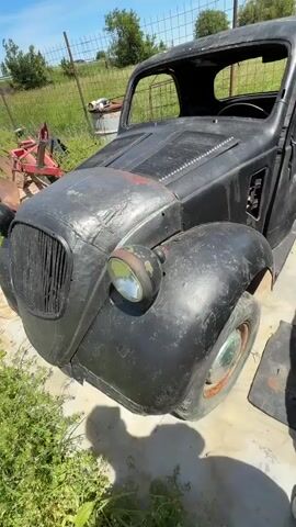 The video shows a vintage car in disrepair. The car body is black and rusty with missing panels and interior parts. The steering wheel is intact, but the rest of the interior is stripped down and rusty. The car is surrounded by grass, a fence, and other vehicles in the background, suggesting it is in a rural or storage area. The weather appears sunny.