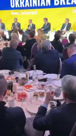 The video shows a room during the "Ukrainian Breakfast" event in Davos, Switzerland. A panel of men in suits sits on a stage behind a long table of seated attendees. The audience, mostly people in business attire, face the stage while eating and listening to the speakers. On the tables are plates with food, water bottles, glasses, coffee cups, and pitchers of milk. The backdrop features a yellow and blue design with the event title in large letters. The focus shifts between the panel and the audience.