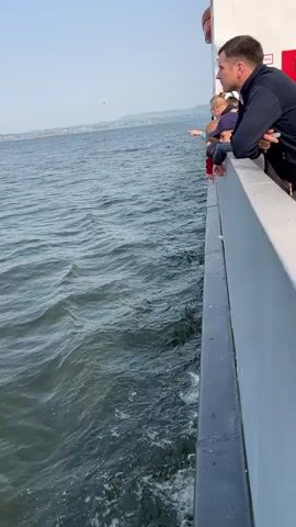 The video shows a view from the side of a boat, looking out at the water. Several people are leaning over the railing on the right side of the frame. A man in a dark jacket is looking out at the water, with his arms resting on the railing. A child is also visible, looking out at the water. The water is choppy, with whitecaps. The sky is clear and blue, with a distant shoreline visible in the background.