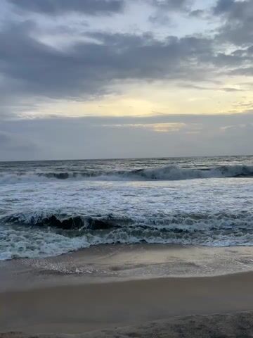 The video shows a wide shot of a beach with rough waves crashing on the shore. The sky is overcast with dramatic clouds, and there is a sliver of yellowish light on the horizon, suggesting either sunrise or sunset. The sand in the foreground is wet and appears to be dark. The waves are frothy and white, with dark green water behind them. The overall impression is one of a stormy or moody seascape.