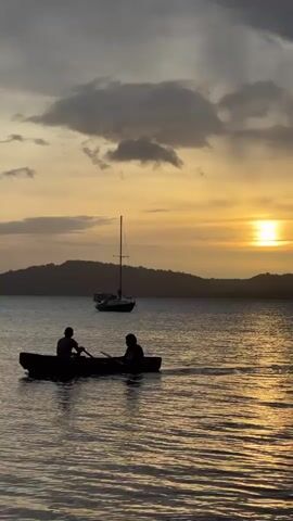 Two people in a rowboat are rowing on the water. A sailboat is anchored in the background. The sun is setting behind a silhouetted mountain range. The sky is filled with dramatic clouds. The water is calm and reflects the colors of the sunset.