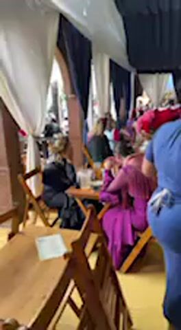 The video shows an indoor or covered outdoor setting, possibly a large tent or decorated hall, serving as a social gathering or dining area in Jerez de la Frontera, Spain. The space is adorned with white and dark blue or black fabric drapes hanging from the ceiling, creating an elegant atmosphere. Numerous people are present, seated at wooden tables on folding wooden chairs.

In the mid-ground, individuals are seen socializing, eating, and drinking. One person in a vibrant purple outfit is seated at a table. The overall scene suggests a busy, lively event. In the foreground, a wooden table with a piece of white paper (text unreadable) is visible. A person wearing a blue t-shirt and glasses walks into the frame, briefly looking towards the camera at the end of the clip. The background is somewhat blurry, but indicates more people and tables. The ambient sound includes chatter and clinking, reinforcing the social nature of the event. The lighting suggests it is daytime.