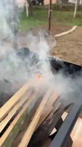 The camera shows wood burning in a black brazier-like container, set outside in a grassy area. The wood catches fire, producing thick white smoke that rises and swirls. A dark object, possibly a handle, appears in the bottom right corner. The background includes blurry trees, fences, and parts of structures, indicating a residential or park-like setting.