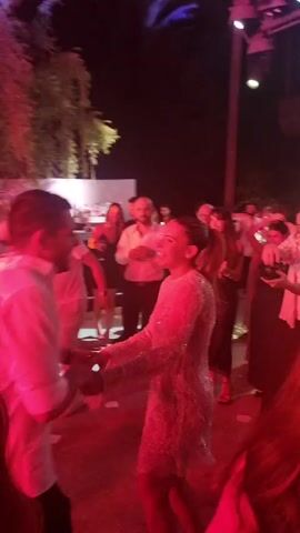 The video shows a group of people dancing at an outdoor event at night. The lighting is dynamic, with flashes of pink, purple, and green lights illuminating the scene. In the foreground, a man in a white shirt is dancing with a woman wearing a sparkling, light-colored dress. Other people are visible in the background, some dancing and some standing around. There appears to be a bar or counter with people behind it. The environment includes trees and some foliage, suggesting an outdoor venue. The atmosphere seems festive and celebratory.