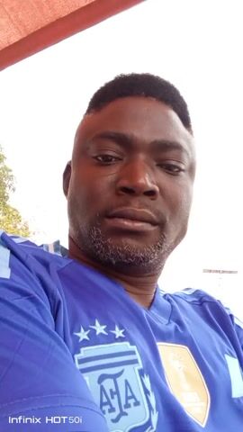 The video shows a close-up shot of a man wearing a blue soccer jersey with the Argentina team logo on it. He has a dark complexion, short black hair, and a short beard with some gray hairs. He is looking directly at the camera with a neutral expression. There is a small scar visible near his left eye. The background is bright and slightly out of focus, showing some trees and a building. The video is taken from a low angle, looking up at the man. The text "Infinix HOT 50i" is visible at the bottom left corner of the screen, indicating the device used to record the video. There is a red awning or roof visible at the top of the frame.